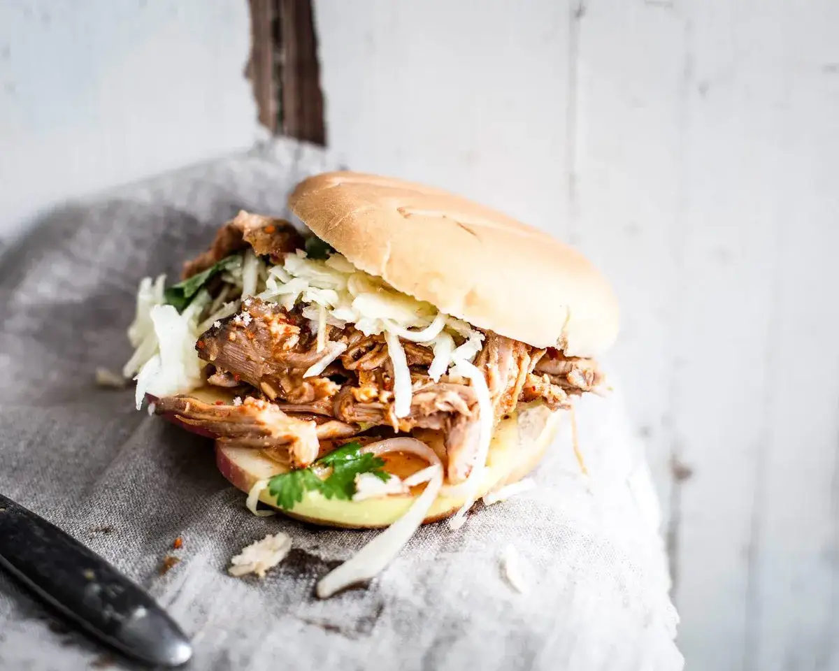Saftiges, gezupftes Fleisch mit Krautsalat und K&auml;se in einem Br&ouml;tchen, serviert auf einem grauen Tuch.