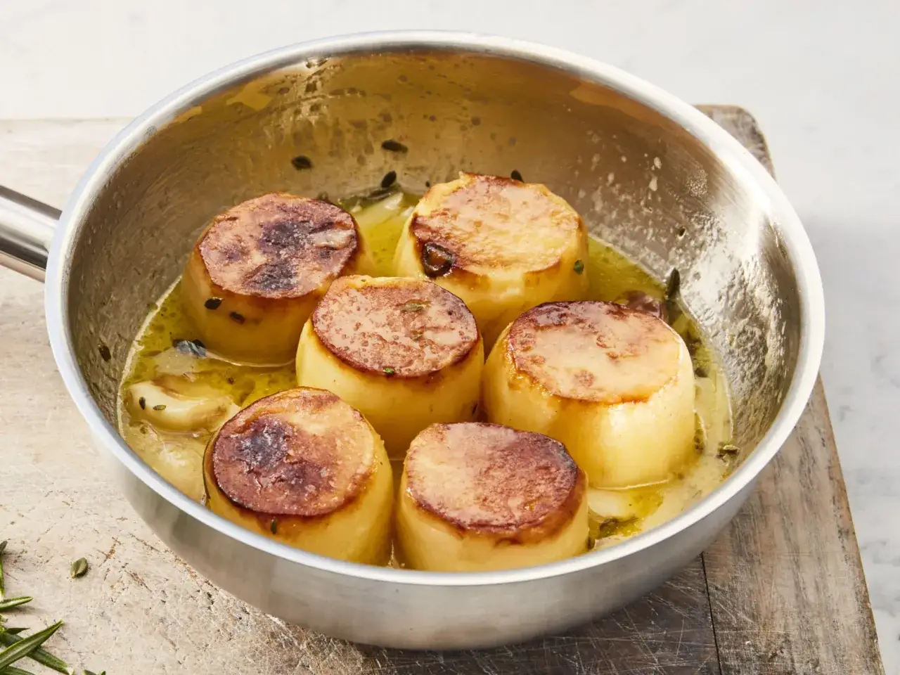 Sechs goldbraune Fondantkartoffeln in einer Pfanne mit Butter und Kräutern, bereit zum Servieren.