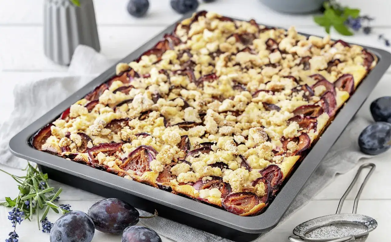 Frisch gebackener Zwetschgenkuchen mit Streuseln in einer rechteckigen Form, garniert mit frischen Zwetschgen und Kr&auml;utern.