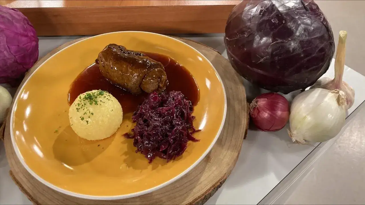 Rinderrouladen mit Rotkohl und Kl&ouml;&szlig;en auf gelbem Teller, daneben frischer Rotkohl, Zwiebeln und Knoblauch.
