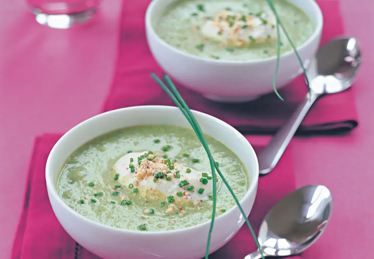 Zwei Schalen cremiger Brokkolisuppe, garniert mit einem Klecks Sahne, Schnittlauch und Nüssen, stehen auf einem pinken Tuch.