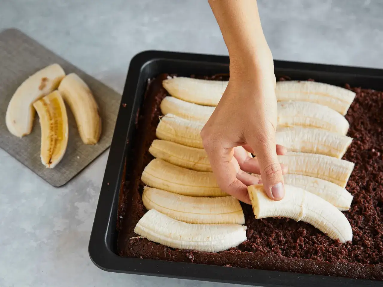 Eine Hand legt Bananenstücke auf einen Schokoladenboden im Blech. Der Maulwurfkuchen vom Blech wird vorbereitet.