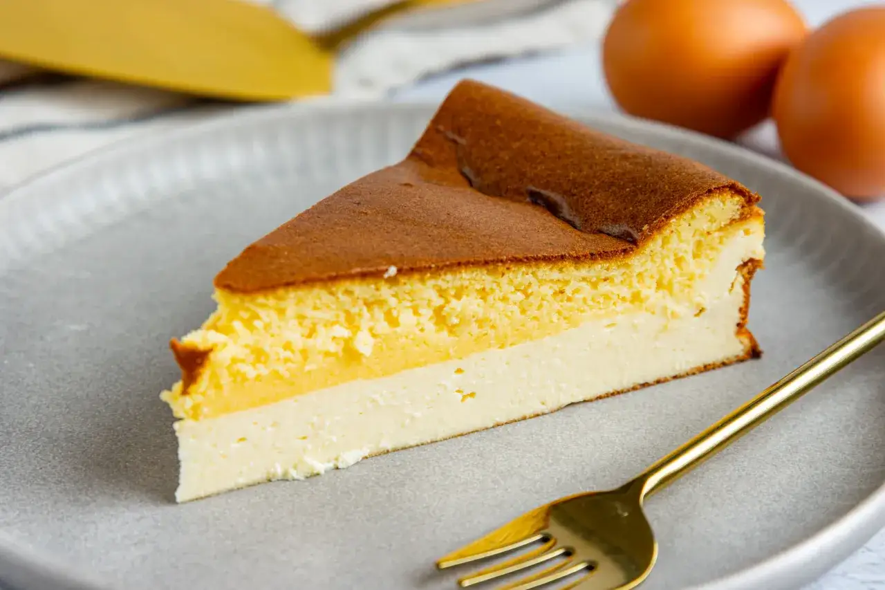 Ein St&uuml;ck Eierschecke auf einem grauen Teller, daneben ein goldener L&ouml;ffel und zwei Eier. Dieses Rezept ist ein Genuss!