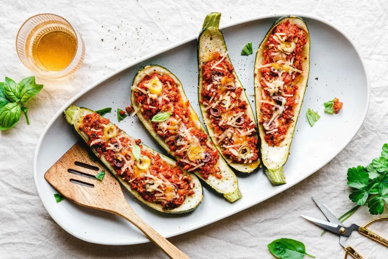 Vier gef&uuml;llte Zucchini-Schiffchen mit Hackfleisch, Tomaten und K&auml;se, garniert mit Basilikum. Daneben ein Glas Wein und frische Kr&auml;uter.