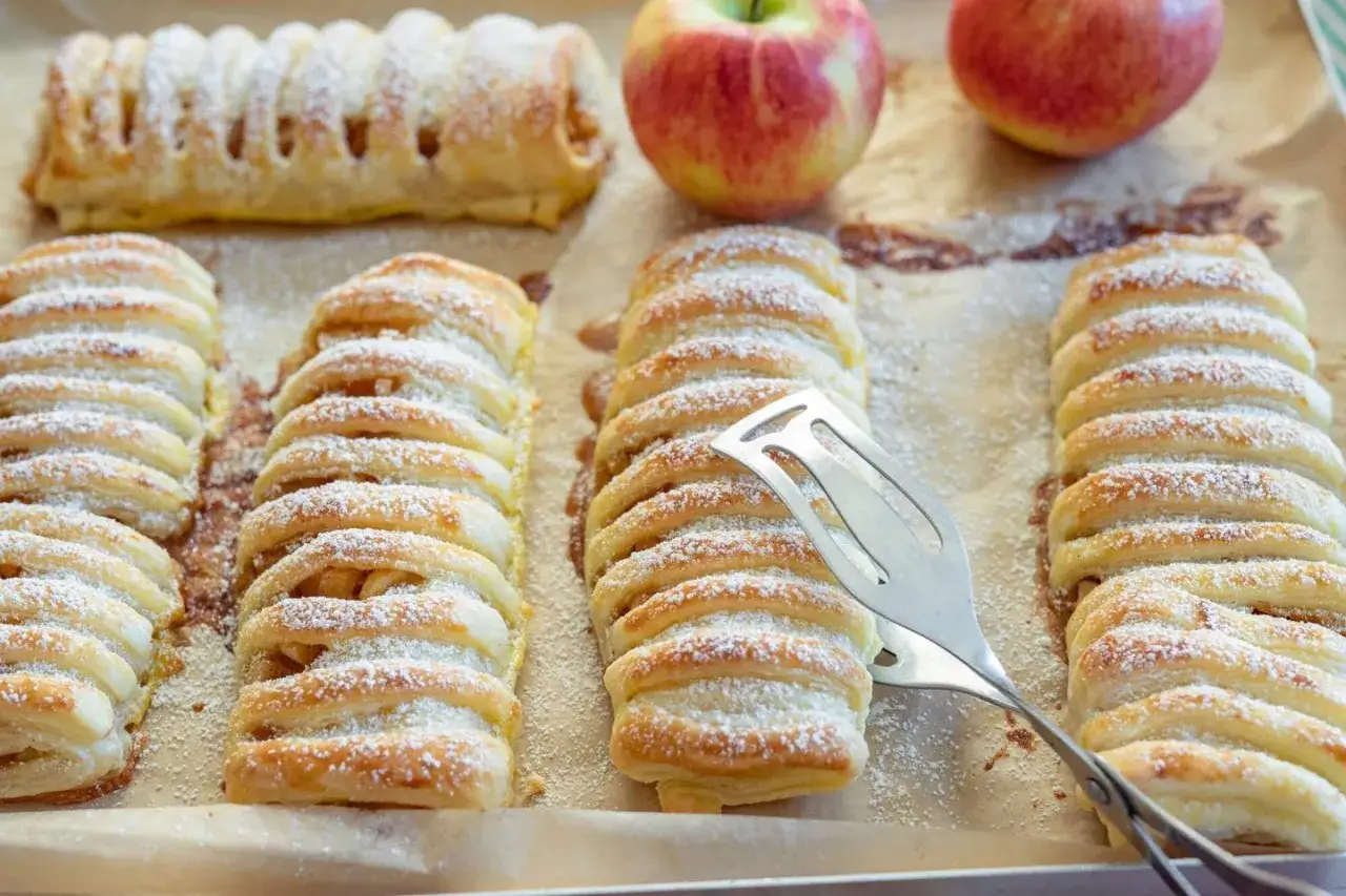Frisch gebackene Apfeltaschen, best&auml;ubt mit Puderzucker, warten auf das Apfeltaschen Rezept. Ein Apfel liegt daneben.