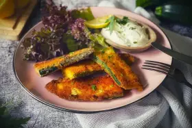 Knuspriges Zucchini-Schnitzel auf rosa Teller, serviert mit Dip, Salat und Zitrone. Ein Genuss für jeden Tag!
