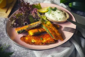 Knuspriges Zucchini-Schnitzel auf rosa Teller, serviert mit Dip, Salat und Zitrone. Ein Genuss für jeden Tag!