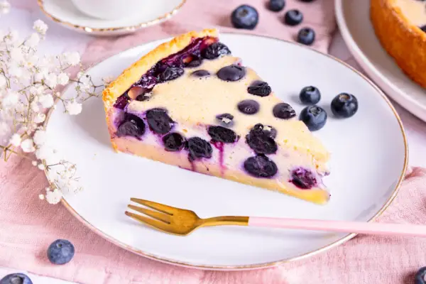 Ein Stück Heidelbeerkuchen Landfrauenart mit saftigen Blaubeeren und cremigem Guss auf einem Teller, daneben eine goldene Gabel und frische Beeren.